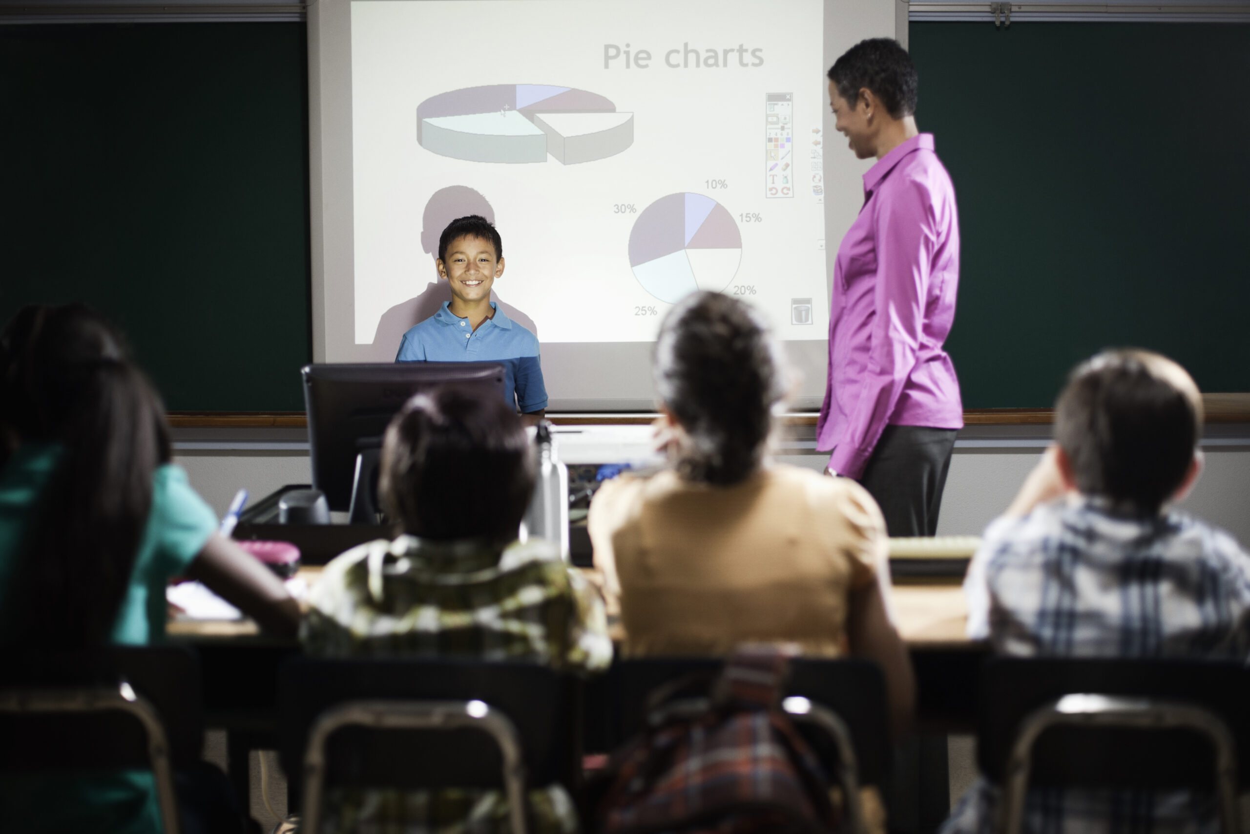 A boy standing at a podium in front of a screen showing pie charts to the class.